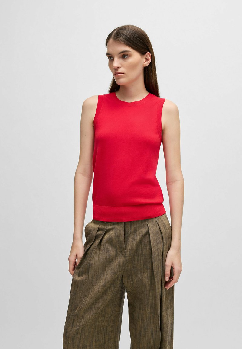 Top senza maniche di maglia rossa con colletto tondo e orlo a coste, abbinato a pantaloni bronze a vita alta con pieghe e un motivo testurizzato.