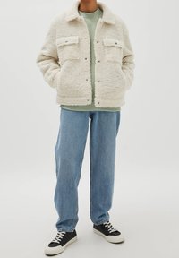 Faux fur cremet jakke med to frontlommer, krave og trykknapper, lagd over en grøn skjorte og parret med lyse jeans og sorte sneakers.