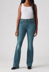 Jeans acampanados azules de mezclilla, con una cintura alta, acentos desgastados y bolsillos delanteros, combinados con una camiseta sin mangas blanca y zapatos negros.