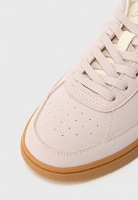 Šviesiai rožiniai zomšiniai sportbačiai su tekstūruota paviršiaus danga, perforacijomis ant piršto dalies, smėlio spalvos raišteliais ir rudu guminiu padu.