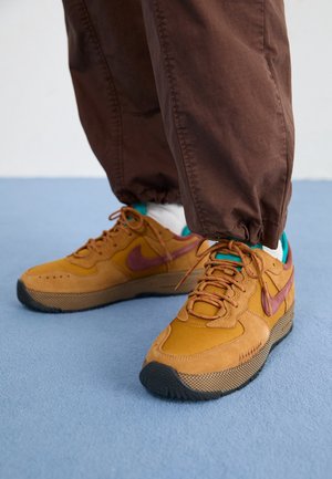 Osoba nosząca brązowe spodnie i musztardowo-żółte buty sportowe z bordowym logo Nike oraz białe skarpetki na niebieskiej podłodze.
