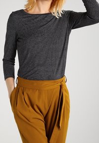 Schwarz-weißer gestreifter Langarmshirt kombiniert mit hoch taillierten, senfgelben Hosen, die eine gebundene Taille und eine plissierte Vorderseite haben.