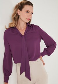 Blusa morada de manga larga, con detalle en el cuello a modo de lazo y puños con fruncidos, hecha de un tejido suave. Combinada con pantalones ajustados de color claro.