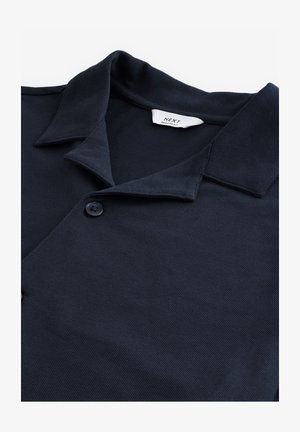 Tumesinine polo särk, mis on valmistatud tekstureeritud kangast ning millel on nööpidega avaus, lühike kaelus ja väike brändi silt kaela juures.