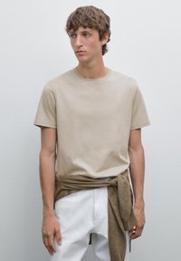 Beige bomulds t-shirt med rund hals, korte ærmer, løs pasform, parret med hvide bukser og en brun sweater bundet om livet.