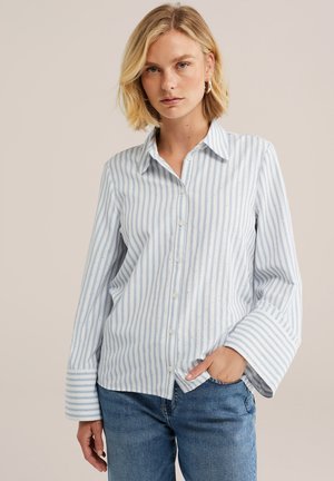 Blonde Frau trägt ein weißes und hellblau gestreiftes Hemd mit breiten Manschetten und blaue Jeans, steht mit einer Hand in der Tasche.