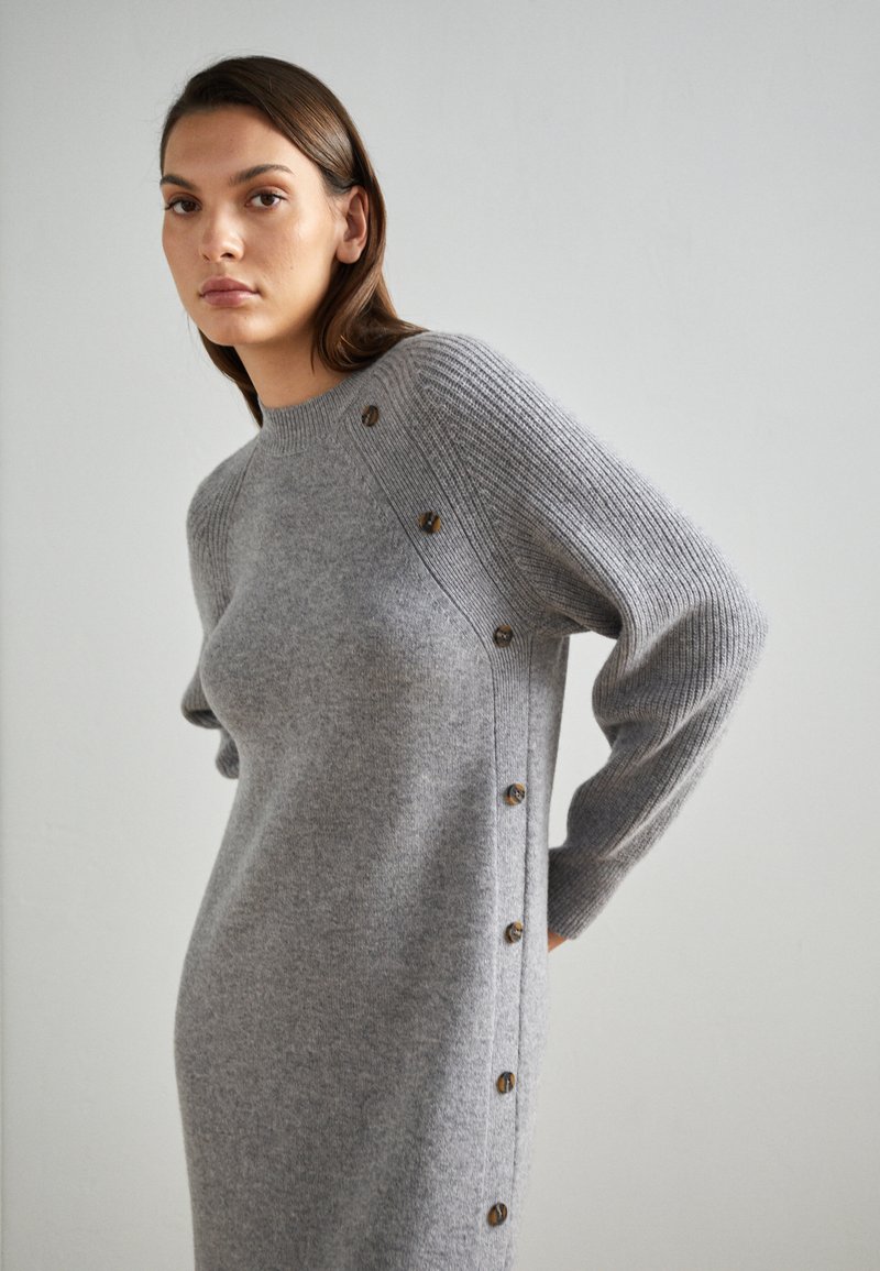Kobieta w dopasowanej szarej sukience dzianinowej z guzikami na ramieniu i boku, stojąca na tle w jasnym kolorze.