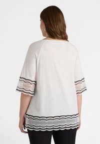 Weißes Oberteil mit kurzen Ärmeln, das mit schwarzer und weißer Spitzenborte an den Ärmeln und dem Saum verziert ist und einen strukturierten, geschichteten Look bietet.
