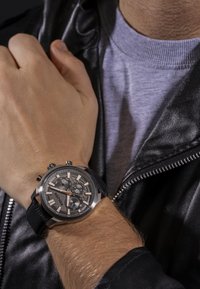 Zegarek chronograf z srebrową kopertą, szarym cyferblatem, różowo złotymi akcentami i czarnym skórzanym paskiem, prezentowany na nadgarstku. Widoczna teksturowana, ciemna skórzana kurtka.