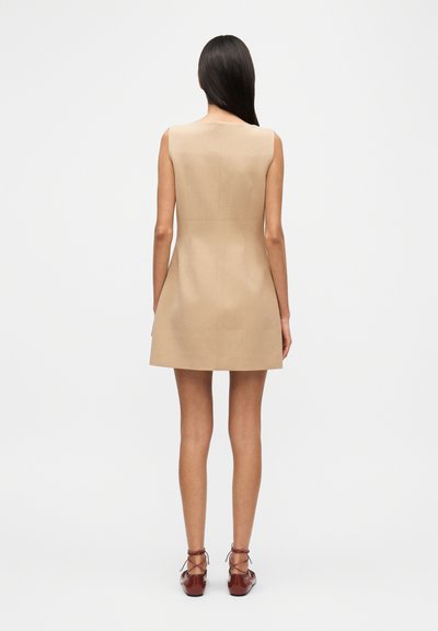 Beigefarbenes ärmelloses Kleid mit tailliertem Oberteil und ausgestelltem Rock, aus glattem Stoff gefertigt. Kombiniert mit braunen Schnürschuhen. Minimalistisches Design.