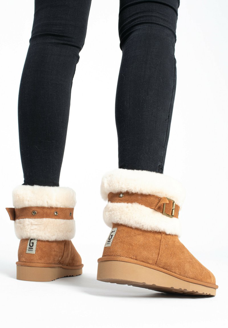 Braune Wildleder-Ankle-Boots mit einem weichen cremefarbenen Fellfutter und -umschlag, versehen mit einer dekorativen Schnalle und einem Logo an der Seite, vor einem weißen Hintergrund.