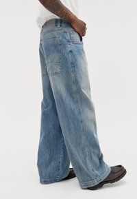 Weite blaue Denim-Jeans mit einer ausgewaschenen Optik, zwei Gesäßtaschen und einer geraden Silhouette, kombiniert mit dunkelbraunen Lederschuhen.