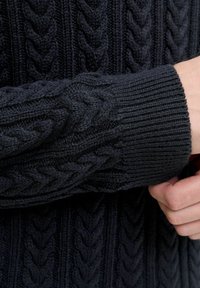Granatowy sweter z dzianiny o teksturowanym splocie w warkocze; ściągacze przy mankietach i dolnej krawędzi zapewniają dopasowanie; materiał wydaje się miękki i gruby.