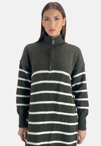 Abito in maglia verde scuro con strisce orizzontali bianche, colletto alto e zip a un quarto. Tessuto morbido e vestibilità rilassata.