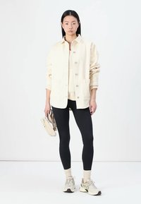 Veste en maille côtelée crème avec de grands boutons, portée avec un legging noir et des baskets claires. Le modèle tient un sac assorti, posé contre un fond uni.