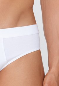 Culottes en coton blanc avec une texture lisse, présentant une large ceinture et une coupe ajustée, partiellement visibles sur une peau bronzée.