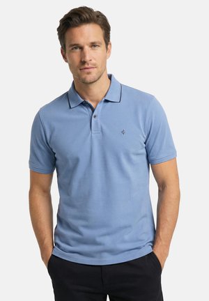 Mann trägt hellblaues Kurzarm-Poloshirt mit Kragen und Knöpfen, steht mit den Händen in den Taschen vor einfarbigem Hintergrund.