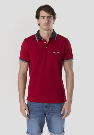 Camisa polo roja con tela texturizada, cuello y detalles en las mangas en azul marino contrastante, y un pequeño logo en el pecho. Combinada con pantalones cortos de mezclilla.