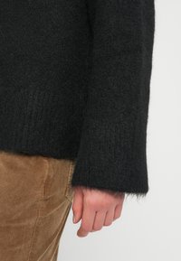 Main reposant sur le côté, portant un pull noir à texture duveteuse et un pantalon en velours côtelé marron, sur un fond clair uni.