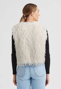 Cremfarbene Kunstpelzweste mit einer strukturierten, flauschigen Oberfläche, die ein kurzes Design und einen geschichteten Look aufweist, über einem schwarzen Langarmshirt und hellblauen Jeans.