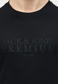 Μαύρο μπλουζάκι με στρογγυλή λαιμόκοψη και ανάγλυκο μαύρο κείμενο "JACK & JONES PREMIUM EST. 1990" στο στήθος, που φοριέται από ένα άτομο.