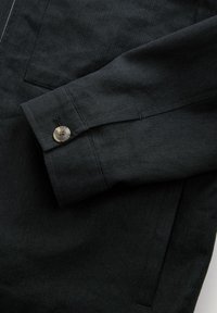 Black fabric jacket with a textured finish, featuring a button cuff and a forward-angled pocket. The material is durable and woven.