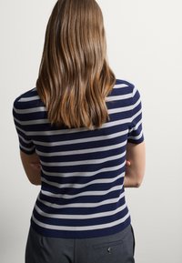 Maglione a righe blu navy e grigio chiaro, realizzato in maglia con maniche corte, design aderente e una consistenza liscia, indossato con pantaloni grigi.
