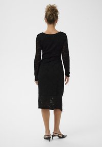 Crna čipkasta haljina s dugim rukavima, prilagođenim siluetom i duljinom do sredine tele. Ima valoviti rub i prozirnu teksturu. Visoke potpetice su vidljive ispod.