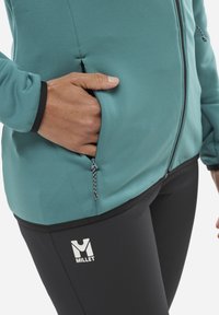 Türkiser Fleecejacke mit schwarzen Akzenten und seitlichen Taschen, kombiniert mit schwarzen, figurbetonten Hosen mit einem weißen Logo. Glattes, dehnbares Material.