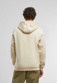 Hoodie beige con un ampio cappuccio morbido, polsini a coste e tasca a marsupio. La parte posteriore presenta un tessuto liscio e testurizzato.