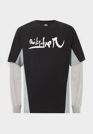 Νέος άνδρας με μαύρο και γκρι μακρυμάνικο T-shirt σε στρώσεις με λευκό γραφικό κείμενο, στέκεται μπροστά σε ένα απλό φωτεινό φόντο.