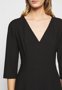 Femme portant une robe noire à col en V avec des manches trois-quarts, montrée de l'épaule jusqu'au milieu du torse sur un fond uni.