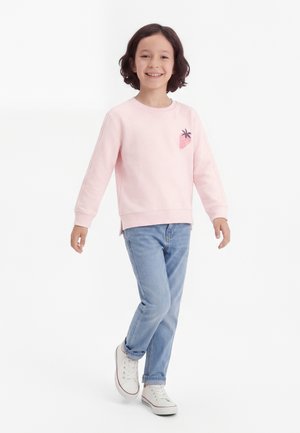 Smaidīgs bērns, kurš staigā, valkājot gaiši rozā džemperi ar zemeņu attēlu, zilas džinsu bikses un baltas sporta kurpes uz baltas fona.