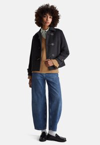 Dunkelgrauer Woll-Pea-Coat mit Knebelverschlüssen über einem beige gerippten Pullover und blauen, weit geschnittenen Jeans, kombiniert mit schwarzen Loafers und weißen Socken.