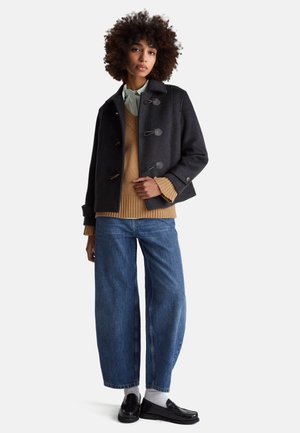 Dunkelgrauer Woll-Pea-Coat mit Knebelverschlüssen über einem beige gerippten Pullover und blauen, weit geschnittenen Jeans, kombiniert mit schwarzen Loafers und weißen Socken.