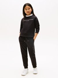 Μαύρη φούτερ μπλούζα με λευκό λογότυπο "TOMMY HILFIGER", σε συνδυασμό με μαύρες φόρμες που διαθέτουν λογότυπα. Λευκά sneakers ολοκληρώνουν το ανάλογο στυλ.