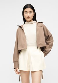 Νέα γυναίκα που φοράει μπεζ κοντό τζάκετ με φερμουάρ πάνω από ένα λευκό τοπ με ψηλό λαιμό και λευκά πλισέ σορτς, ποζάροντας με ουδέτερη έκφραση.