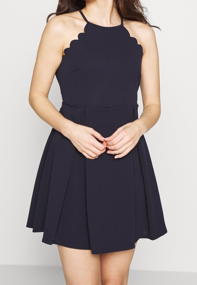 Laivastonsininen halterneck-mekko, jossa on uurrettu kaula-aukko, tiukka vartalo ja liehuva taitettu hame. Valmistettu sileästä, teksturoidusta kankaasta.