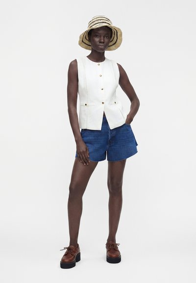 Femme à la peau foncée portant un gilet blanc sans manches boutonné, un short en jean bleu, des chaussures compensées marron et un chapeau beige rayé à large bord.
