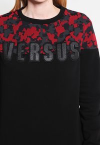 Felpa nera con un motivo camouflage rosso e nero nella parte superiore e la scritta "VERSUS" in un materiale nero testurizzato.