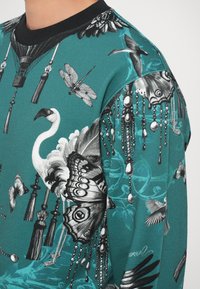 Maglione realizzato in tessuto verde acqua con illustrazioni dettagliate in bianco e nero di uccelli, farfalle e motivi decorativi. Collo a costine nero.