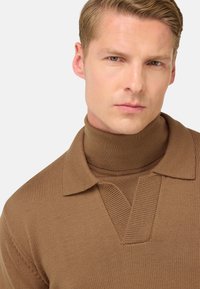 Nahaufnahme eines Mannes, der einen braunen Strickpullover über einem passenden braunen Rollkragenpullover trägt und mit neutralem Ausdruck geradeaus schaut.