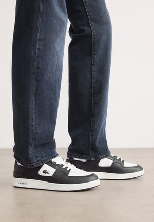 Sorte og hvite Lacoste sneakers med en teksturert skinnoverdel, med snøring og en hvit gummisåle. Brukt med mørke jeans.