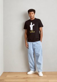 Crna pamučna majica s karikaturom lika koji drži krafnu, s natpisom "The Simpsons" u žutoj boji. Model nosi svijetloplave traperice.