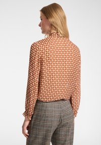 Langärmlige Bluse in Rost mit cremefarbenem geometrischem Muster, mit hohem Kragen und elastischen Bündchen, kombiniert mit grauen karierter Hose.