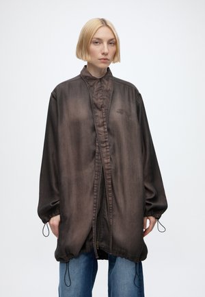 Jeune personne aux cheveux blonds courts portant une veste zippée marron foncé surdimensionnée avec des poignets élastiques et un jean bleu sur un fond uni.