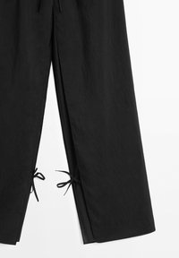 Black pants with wide legs feature slits at the bottom tied with thin black fabric bows on a white background.