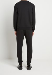 Sudadera negra y pantalones a juego hechos de tela suave. Puños y cintura acanalados; textura suave. Llevado con zapatos negros brillantes.