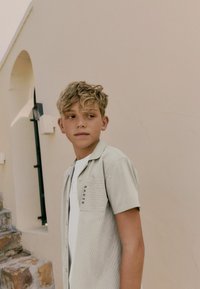 Hellgrünes kurzärmliges Hemd mit einem vertikalen Streifenmuster, ausgestattet mit einer Brusttasche und dem Wort "BAKER" gestickt. Über einem weißen T-Shirt getragen.