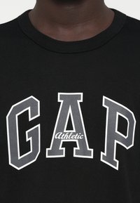 Czarna bawełniana koszulka z dużym szarym napisem "GAP" i mniejszym "Athletic" poniżej, w kontrastowej białej obramówce. Okrągły dekolt.